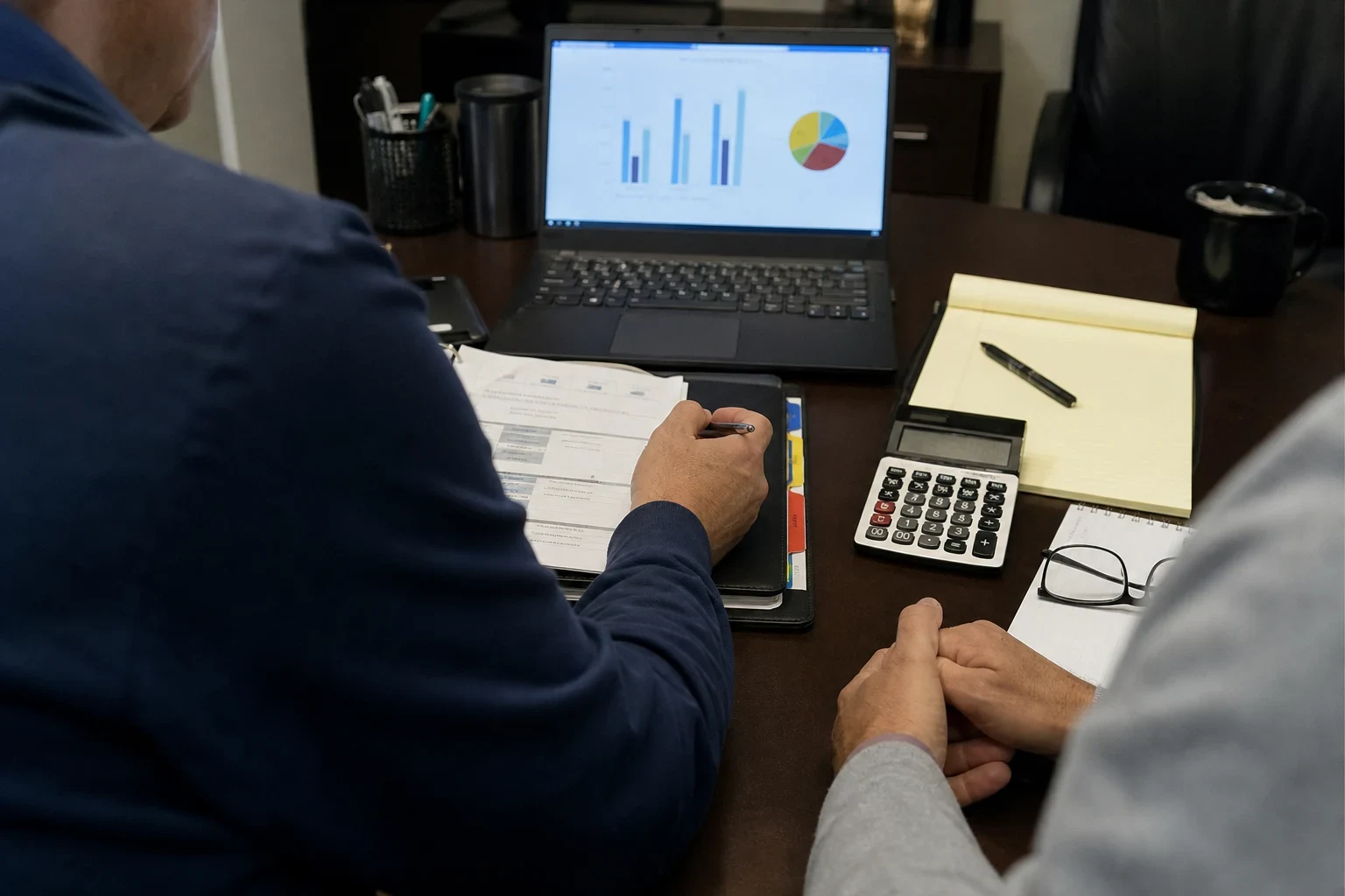 Over-the-shoulder view of two people at an office desk reviewing paperwork beside a laptop showing bar and pie charts, a calculator, legal pad and reading glasses