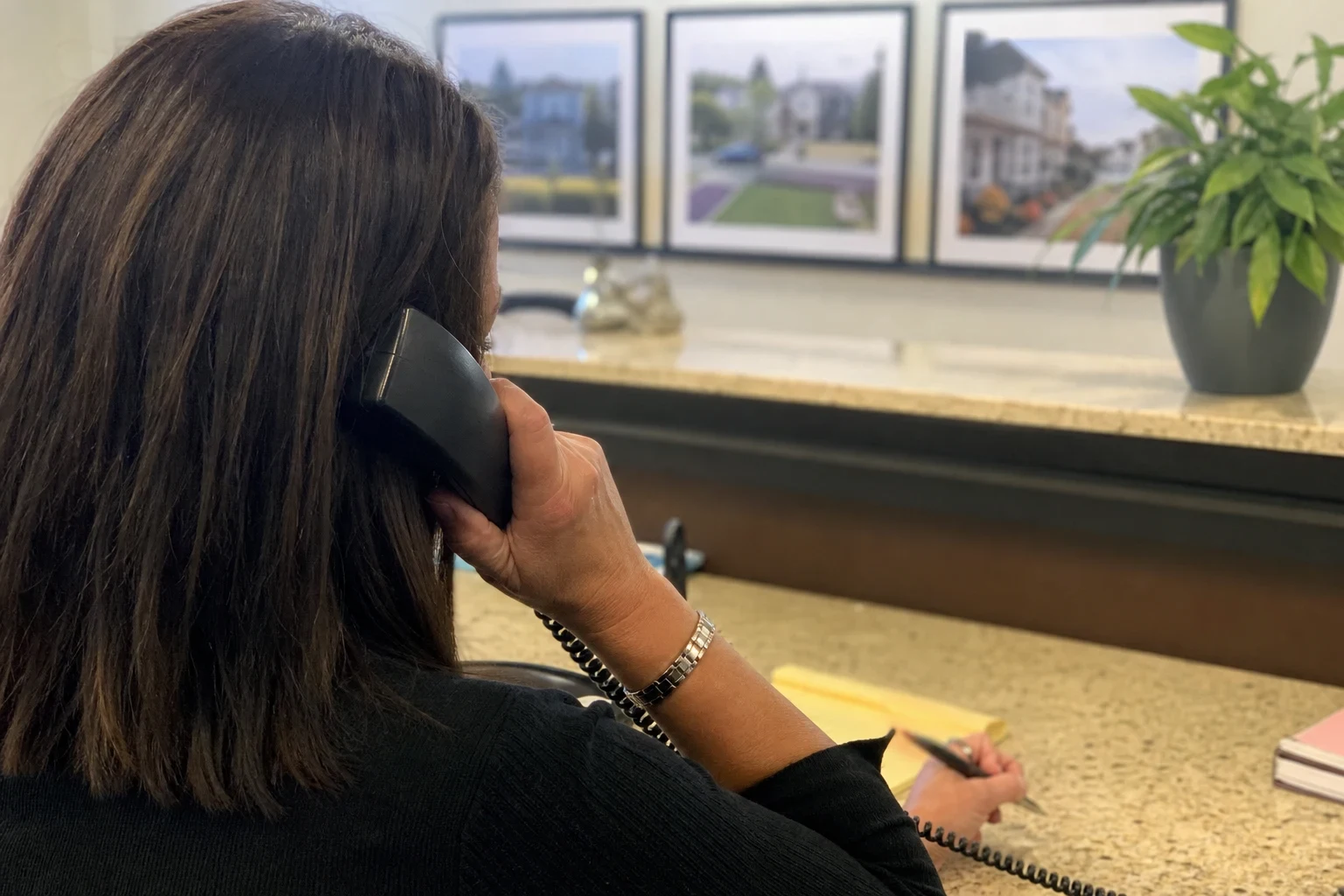 Over-the-shoulder view of an agent on a corded desk phone, taking notes on a yellow notepad at a granite counter, with framed neighborhood photos and a potted plant behind