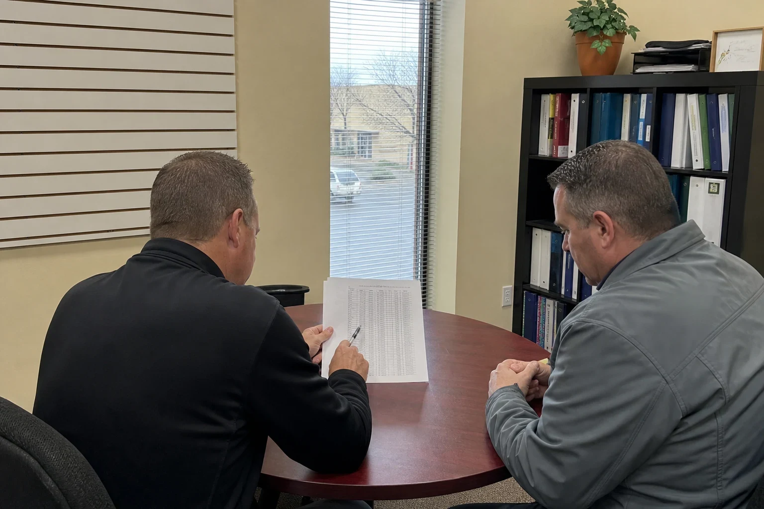 Two men at a small round wood table in an office reviewing a printed page of figures, with a binder bookshelf, slatwall, and a window onto a parking lot