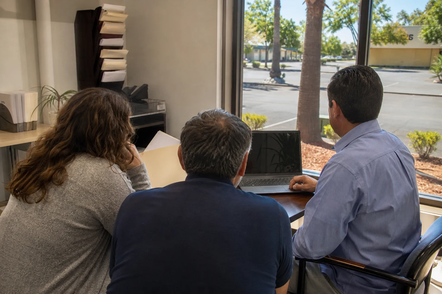 Agent at a laptop with a couple holding a folder at a small table by an office window overlooking palm trees and a sunny parking lot